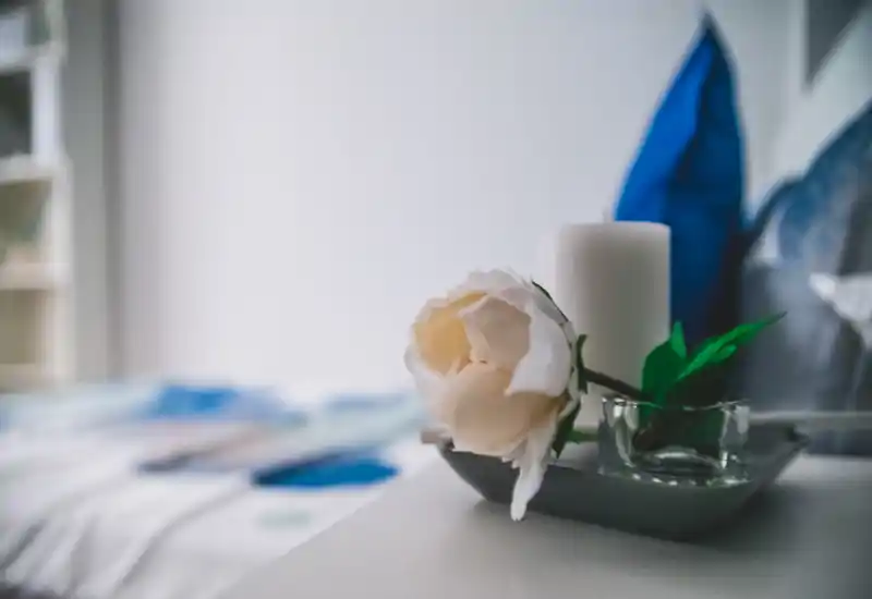 Primo piano di un comodino con candela, fiore decorativo e letto sfocato sullo sfondo — un dettaglio della camera da letto dal look morbido e accogliente.