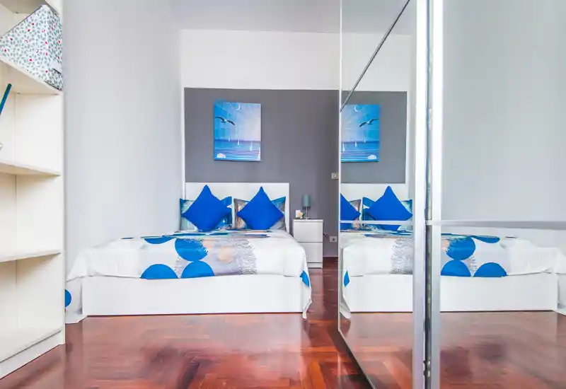 Camera da letto luminosa e moderna con letto bianco, biancheria con accenti blu, cuscini decorativi e armadio con specchio che crea profondità.