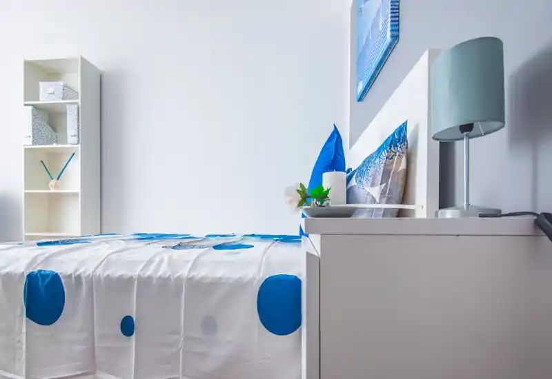 Camera da letto luminosa e ordinata con colori bianco e blu, letto ben visibile con biancheria a fantasia, comodino con lampada e piccola pianta; perfetta per annunci di appartamenti che valorizzano camere minimal e accoglienti.