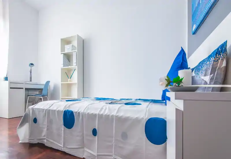 Camera singola luminosa e moderna con biancheria bianca e blu, comodino, scrivania e mensole — stile minimal pulito e ottima luce naturale.