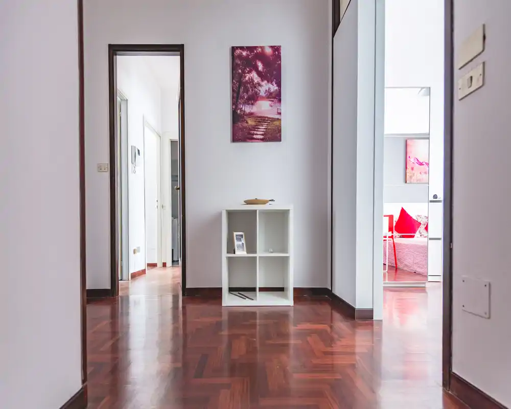 Ingresso luminoso e minimale con pavimento in parquet lucido, un piccolo mobile contenitore e porte che si aprono alle stanze adiacenti — impressione iniziale pulita e accogliente.