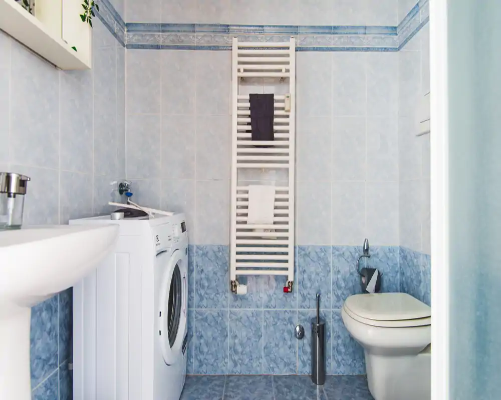 Bagno compatto con piastrelle azzurre, WC, lavabo a colonna e lavatrice a carica frontale; luminoso, pulito e funzionale.