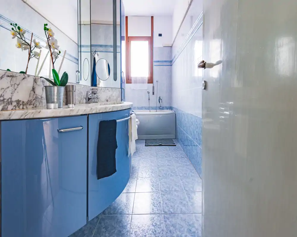 Bagno luminoso e pulito con pavimento e pareti piastrellate azzurre, mobile lavabo curvo con piano in marmo, specchi contenitori e vasca sul fondo.