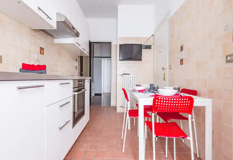 Cucina moderna e luminosa con mobili bianchi, forno incassato e tavolo da pranzo con sedie rosse — spazio funzionale e curato, ideale per affitti brevi.