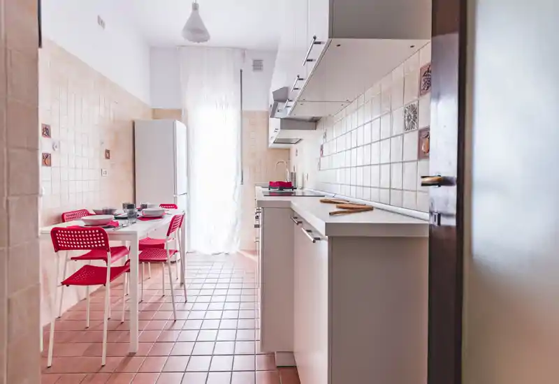 Cucina luminosa e ben curata con pensili bianchi, paraschizzi piastrellato e un piccolo tavolo con sedie rosse — perfetta per annunci di monolocali o bilocali.