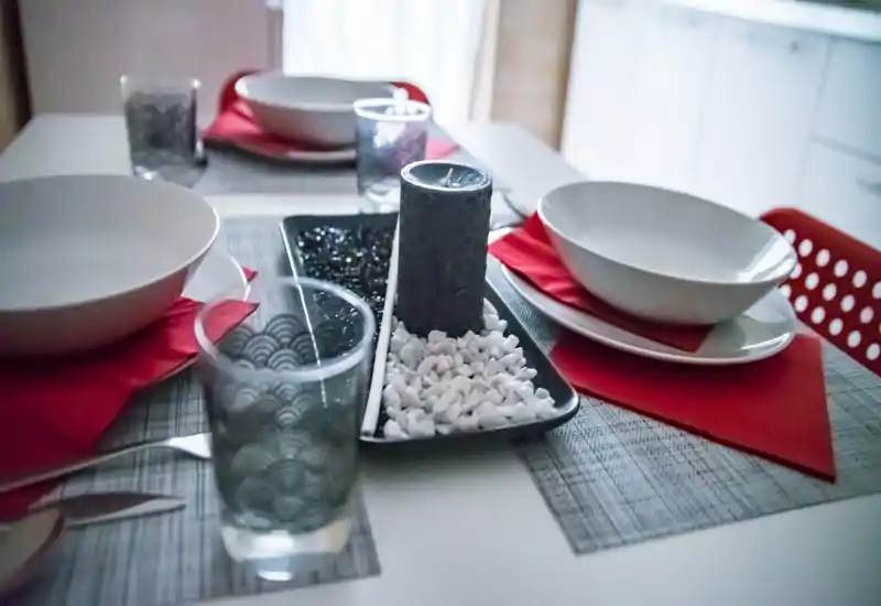 Tavolo da pranzo allestito per quattro con ciotole bianche, tovaglioli rossi e un vassoio decorativo centrale con candela e sassi — angolo pranzo moderno e accogliente.