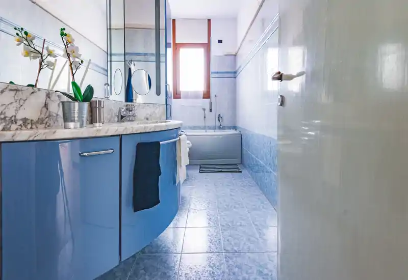 Bagno luminoso e moderno con pavimento e pareti piastrellate azzurre, mobile lavabo curvo con piano in marmo e vasca sotto la finestra.