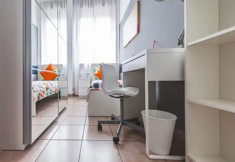 Camera singola moderna e luminosa con scrivania, armadio con specchio e cuscini colorati. La luce naturale dalla finestra crea un ambiente fresco e ordinato, ideale per studenti o ospiti.