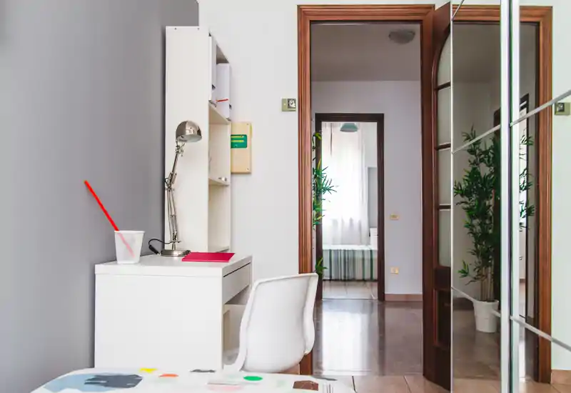 Angolo camera luminoso e ordinato con scrivania bianca e sedia, vista parziale del letto in primo piano e una porta che conduce a un'altra stanza; piante e armadio con specchio aumentano la profondità.