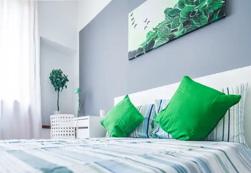 Camera da letto luminosa e moderna con cuscini verdi, biancheria a righe e un quadro sopra la testata. La luce naturale e l’arredo armonioso creano un ambiente fresco e accogliente, perfetto per annunci.