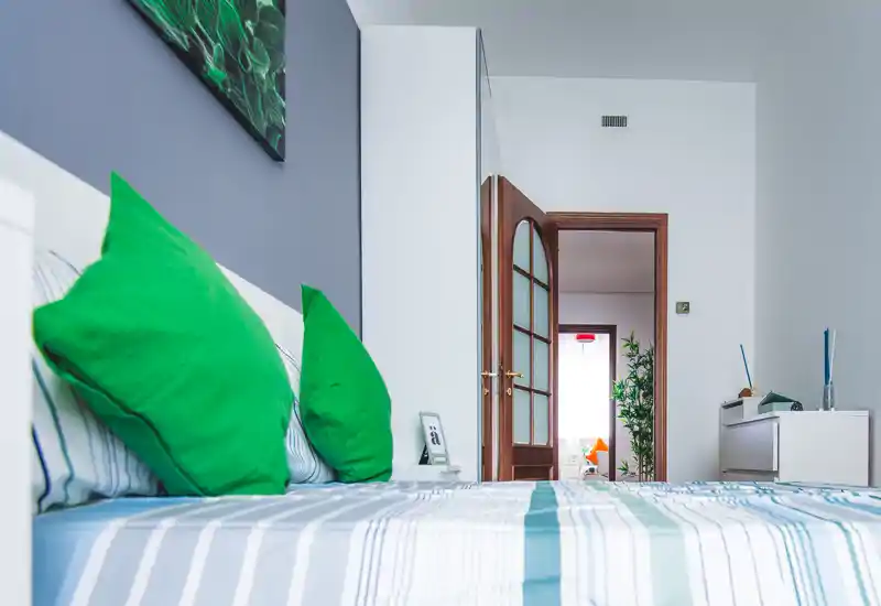 Camera da letto moderna e luminosa con biancheria a righe e cuscini verdi vivaci; design pulito e luce naturale la rendono accogliente.