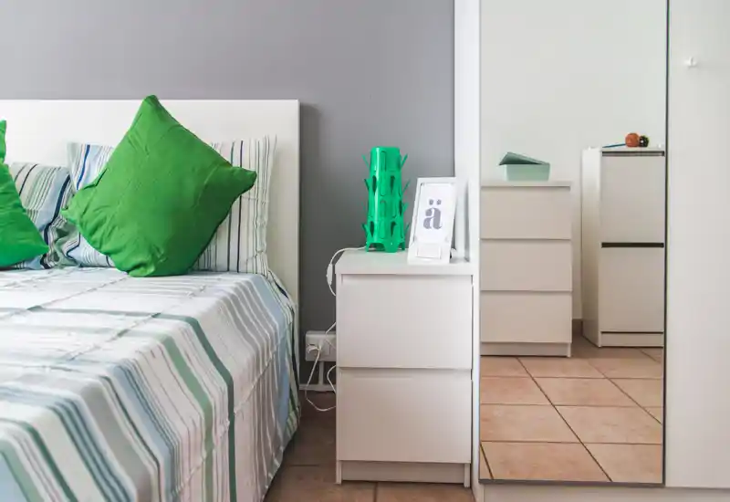 Angolo camera luminoso e moderno con letto a righe, cuscini verdi e comodino bianco accanto a un armadio con specchio — pulito e accogliente.