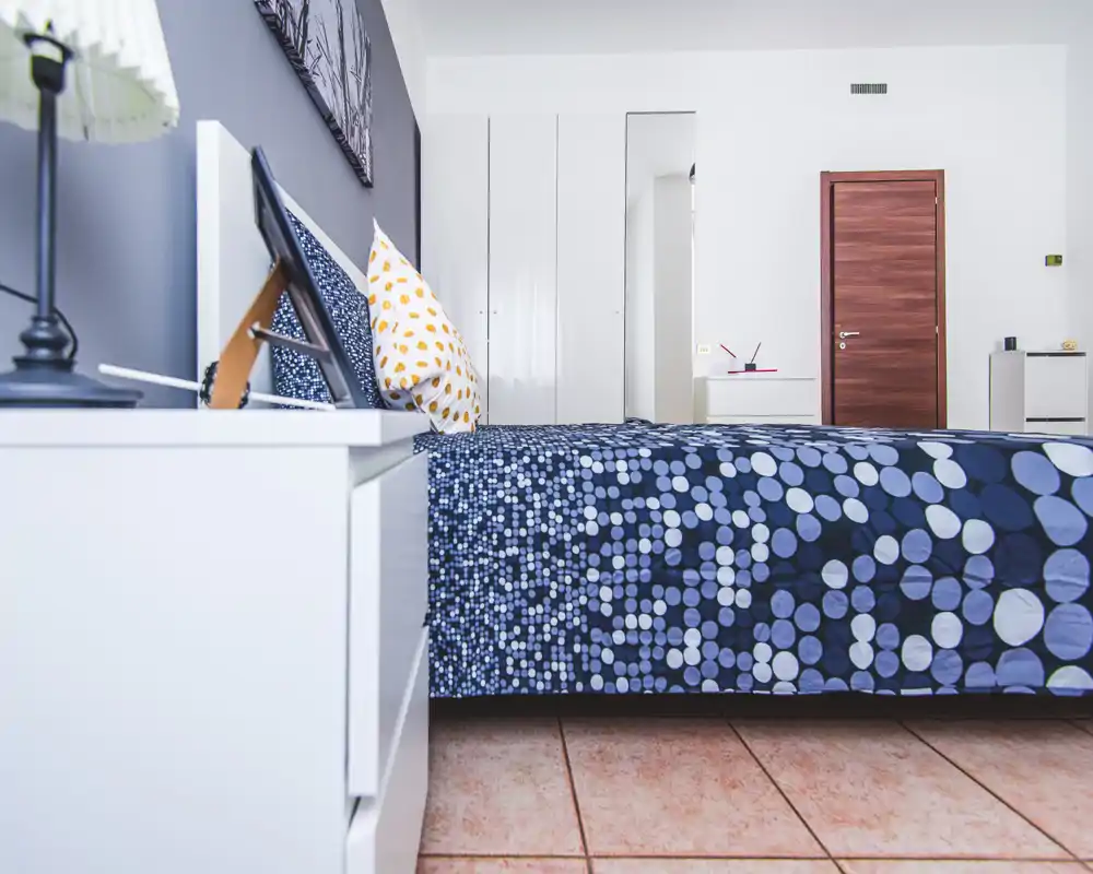 Camera da letto moderna e luminosa con copriletto blu a pois, comodino con lampada e armadio bianco: arredo pulito e minimal.