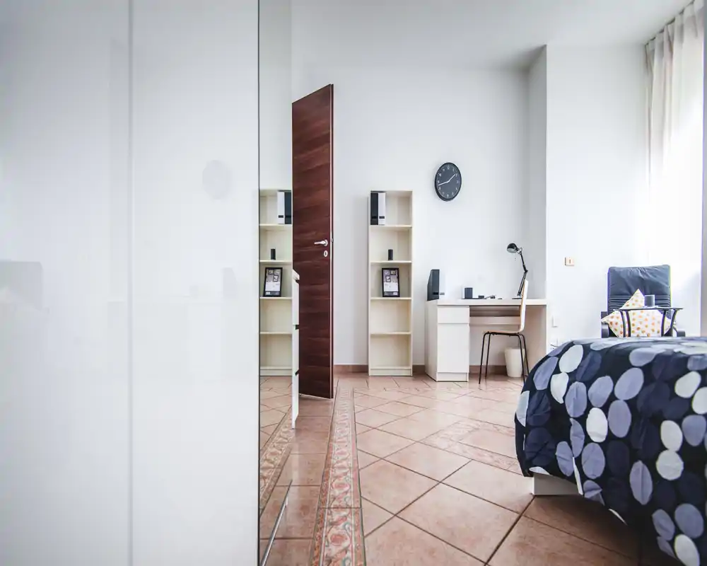 Camera da letto luminosa e minimal con copriletto a motivi, poltrona e scrivania; luce naturale abbondante e stile pulito e moderno.