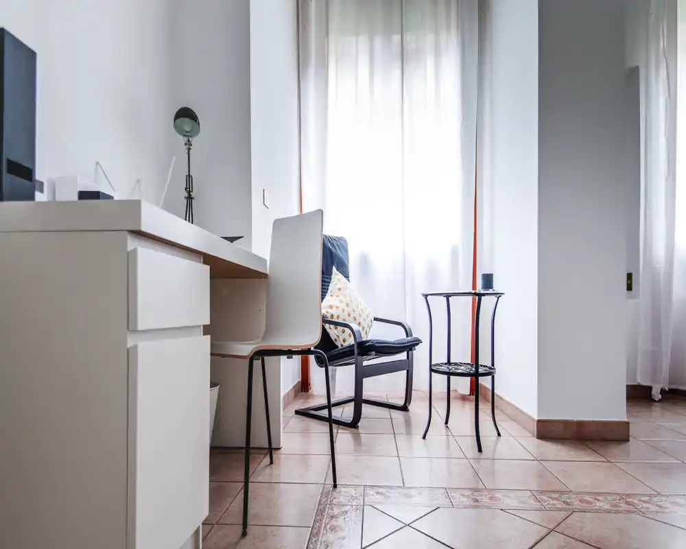 Angolo soggiorno luminoso e minimal con una poltrona comoda, un piccolo tavolino e una scrivania vicino a una grande finestra che diffonde luce naturale.