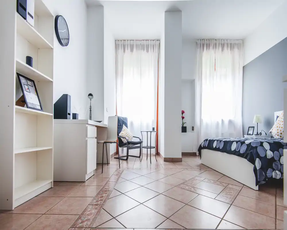 Camera da letto luminosa e moderna con letto singolo, angolo scrivania e poltrona. La luce naturale entra da due grandi finestre; tessili e accessori rendono lo spazio accogliente e funzionale.