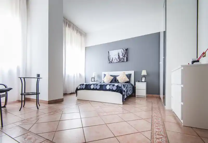 Bright, modern bedroom with a double bed, bedside tables and soft natural light from large windows. Clean lines, neutral gray accent wall and simple decorative pillows create a welcoming, minimal look.