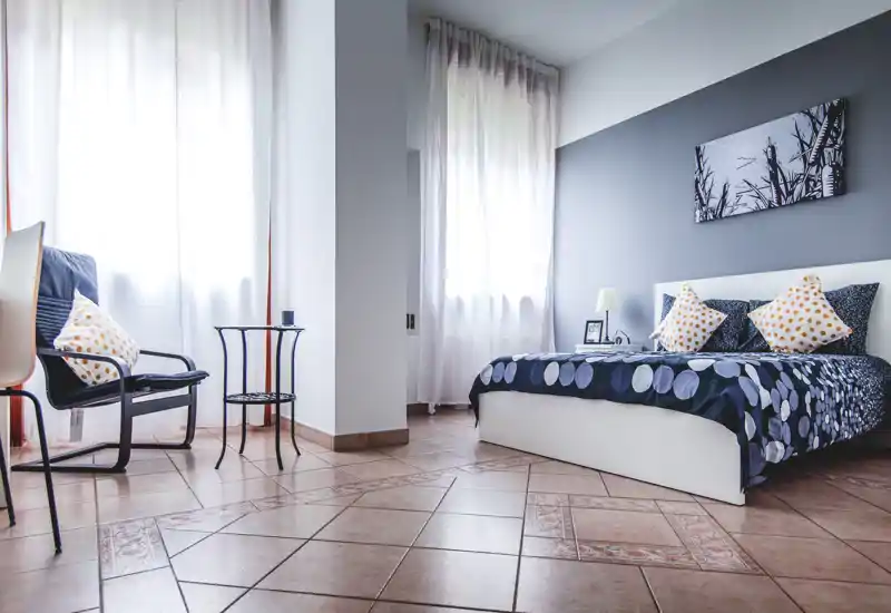 Bright, modern bedroom with a neatly made double bed, patterned bedding and decorative cushions. Large windows with sheer curtains fill the room with natural light and a comfortable sitting area is visible.