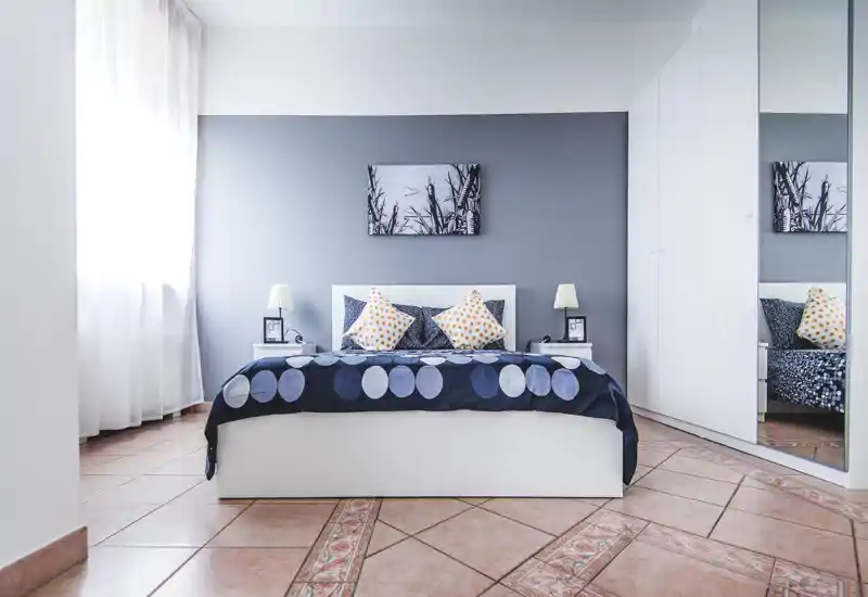 Bright, modern bedroom with a double bed, decorative pillows and bedside lamps; soft natural light from a large window and a mirrored wardrobe enhance the sense of space.
