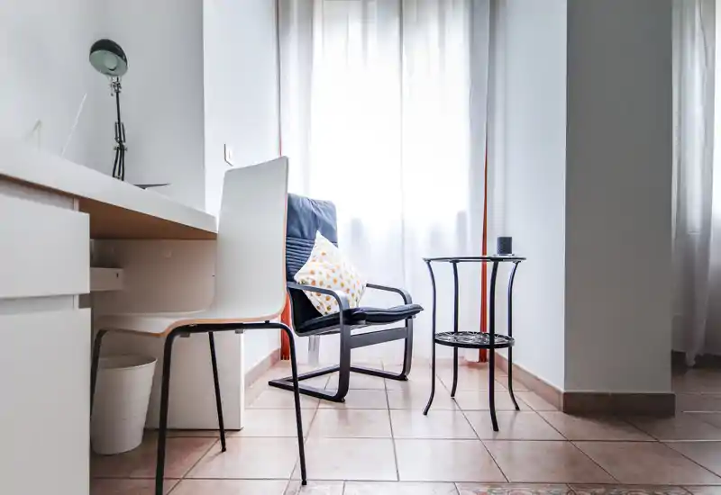 Bright, minimalist living corner with a comfortable armchair, small side table and a desk chair by a sunlit window — ideal for reading or working.