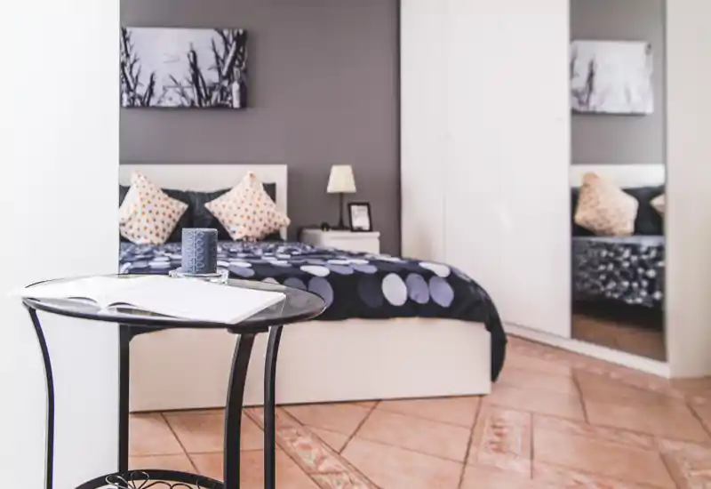 Bright, modern bedroom with a neatly made double bed, patterned cushions and a bedside table with lamp. A mirrored wardrobe and warm floor tiles complete the inviting space.