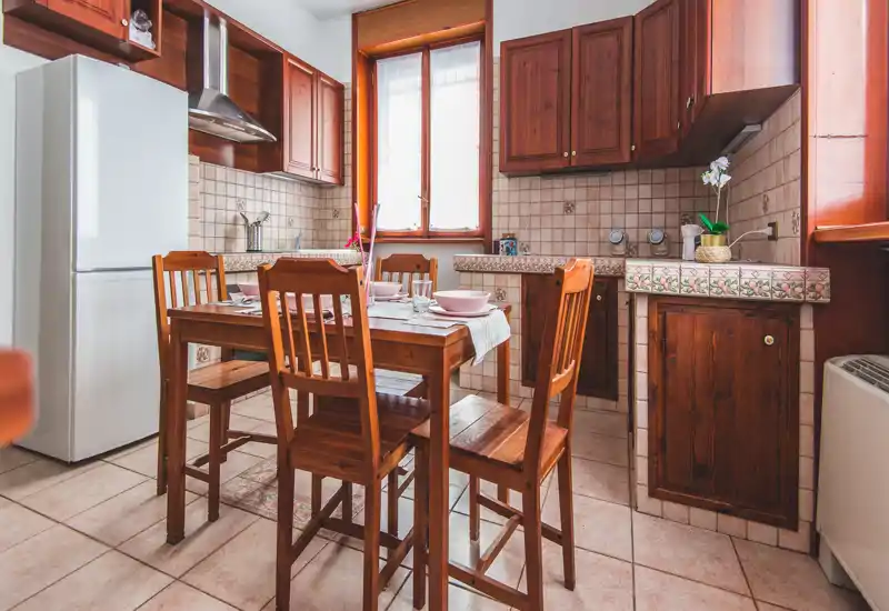 Warm, well-kept kitchen with wooden cabinets and a central dining table set for four. Natural light from a window brightens the tiled counters and decorative backsplash, creating a cozy, homely atmosphere ideal for apartment listings.