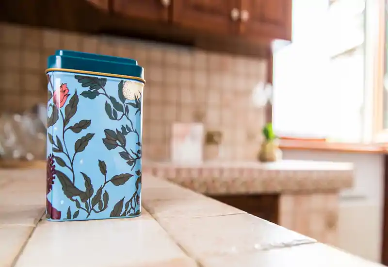 Close-up of a decorative tin on a tiled kitchen counter with warm wooden cabinets and natural window light, highlighting a cozy, lived-in kitchen corner.