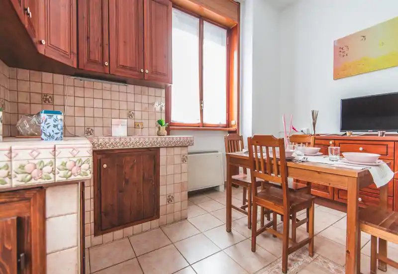 Sunny, traditional kitchen with wooden cabinets and a dining table set for four; warm tiled backsplash and natural light from the window create a welcoming atmosphere.