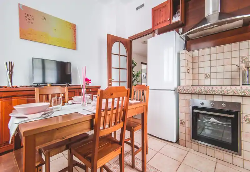 Bright, well-composed kitchen with wooden dining table, chairs and tiled cooking area; fridge and oven visible, ideal for rental photos showcasing a cozy eat-in kitchen.