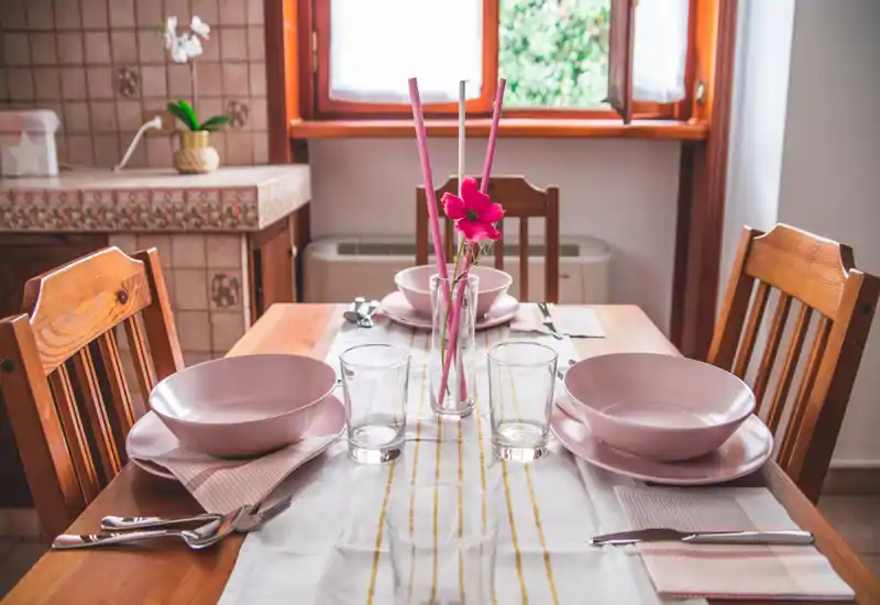 Bright, homely dining area with a wooden table set for four, pink dishware and a floral centerpiece by a sunlit window.