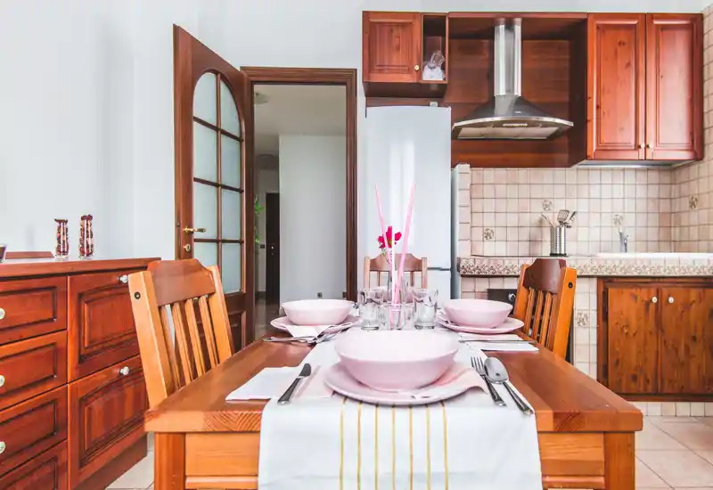 Bright, warmly furnished dining area with a wooden table set for four, matching chairs and a background kitchenette with wooden cabinets.