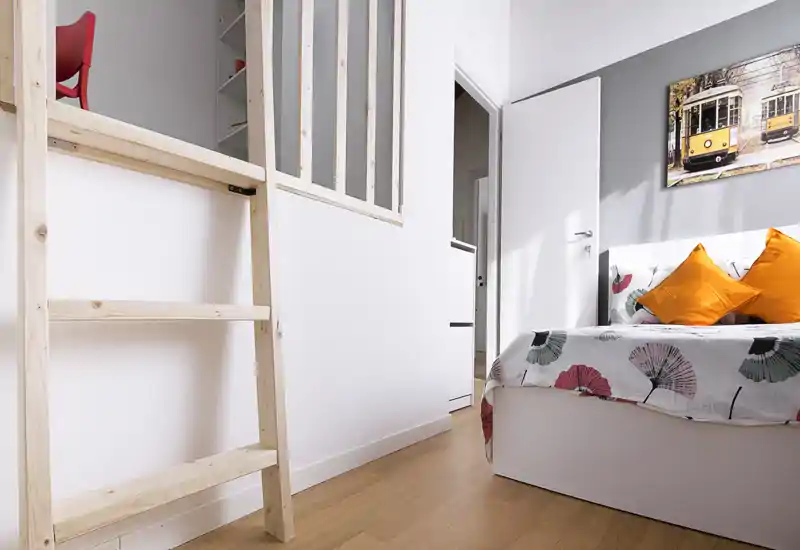Dormitorio moderno y luminoso con ropa de cama estampada y cojines naranjas; suelo de madera clara y detalle de escalera minimalista.