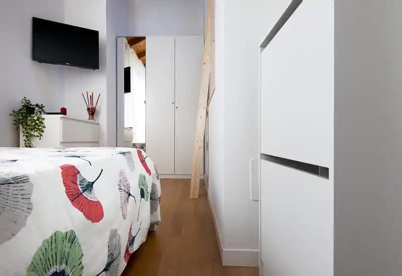 Dormitorio moderno y luminoso con colcha estampada, televisor de pared y armario al fondo. Líneas limpias, suelo de madera y pequeños detalles decorativos como una planta y un jarrón.
