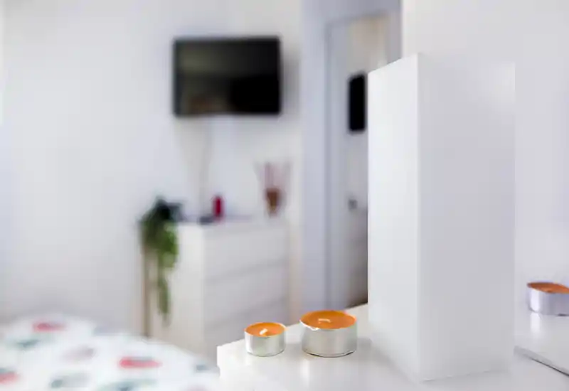 Dormitorio luminoso y minimalista con velas decorativas en primer plano y cama parcialmente visible junto a una cómoda al fondo.