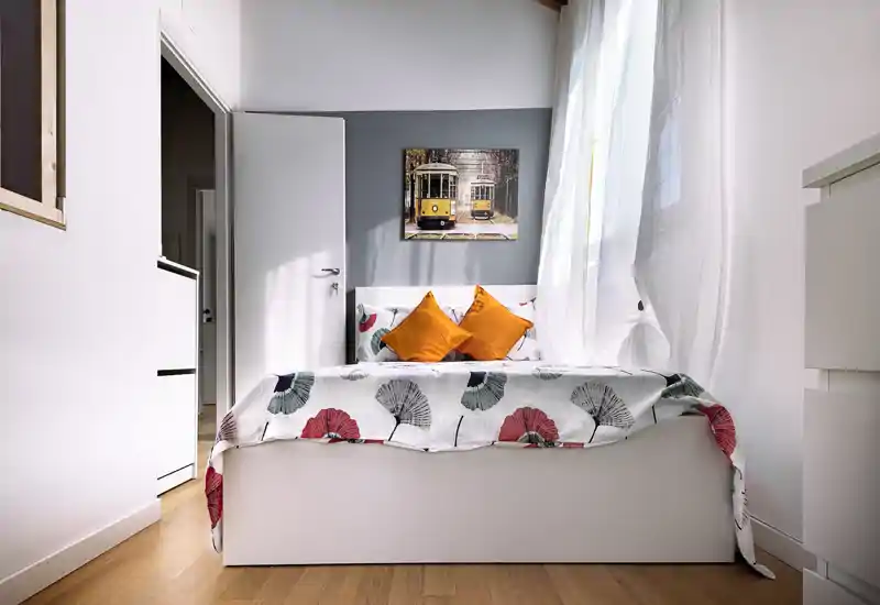 Dormitorio luminoso y bien presentado con ropa de cama estampada y dos cojines naranjas; ventana grande que aporta abundante luz natural, espacio limpio y acogedor.