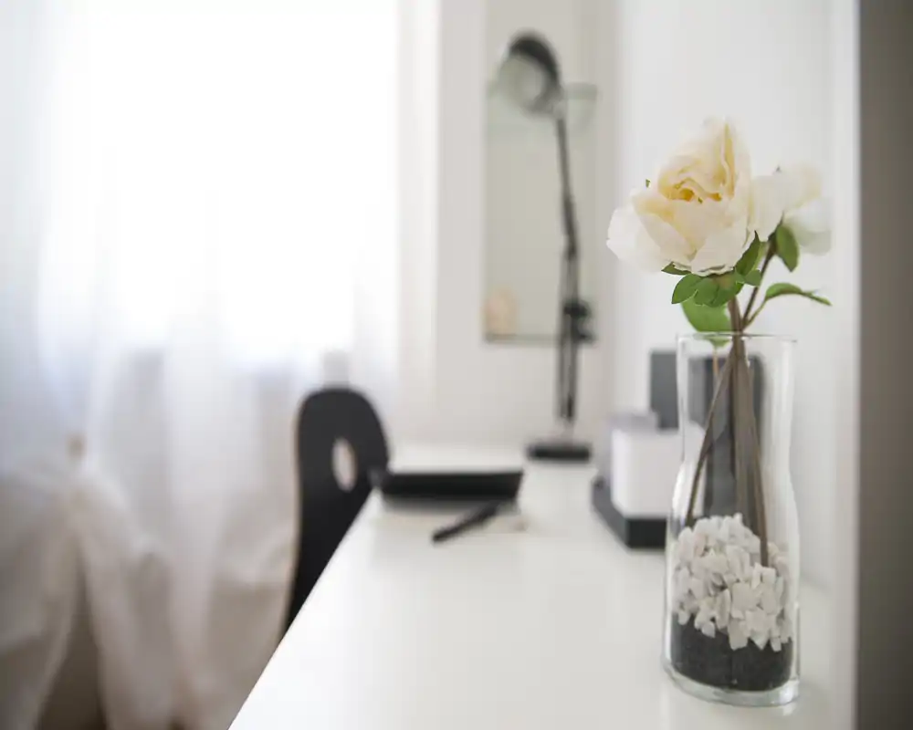 Bright, minimalist living area with a white desk, decorative vase with flowers and a lamp — a calm workspace corner by a curtained window.