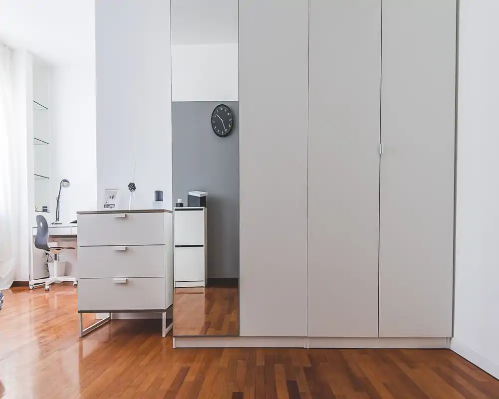 Minimal, modern bedroom corner with large white wardrobes, a mirrored door and a small desk by the window. The wooden floor and bright natural light create a clean, airy atmosphere.