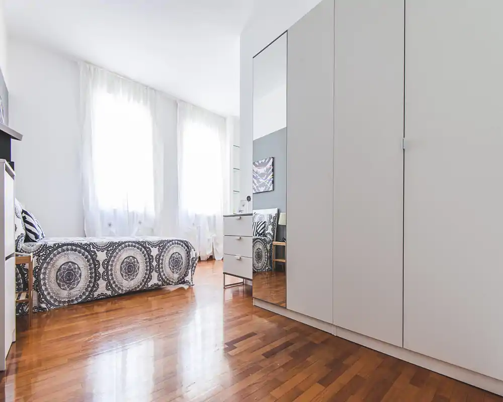 Bright, airy bedroom with a single bed dressed in a patterned cover, large windows with sheer curtains and a mirrored wardrobe that expands the sense of space.