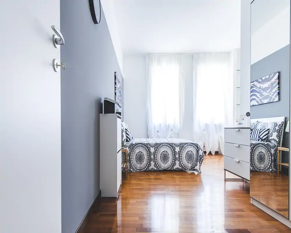 Bright, modern bedroom with a patterned single bed, white curtains and polished wooden floors. The room is freshly styled with minimal furniture and decorative cushions.