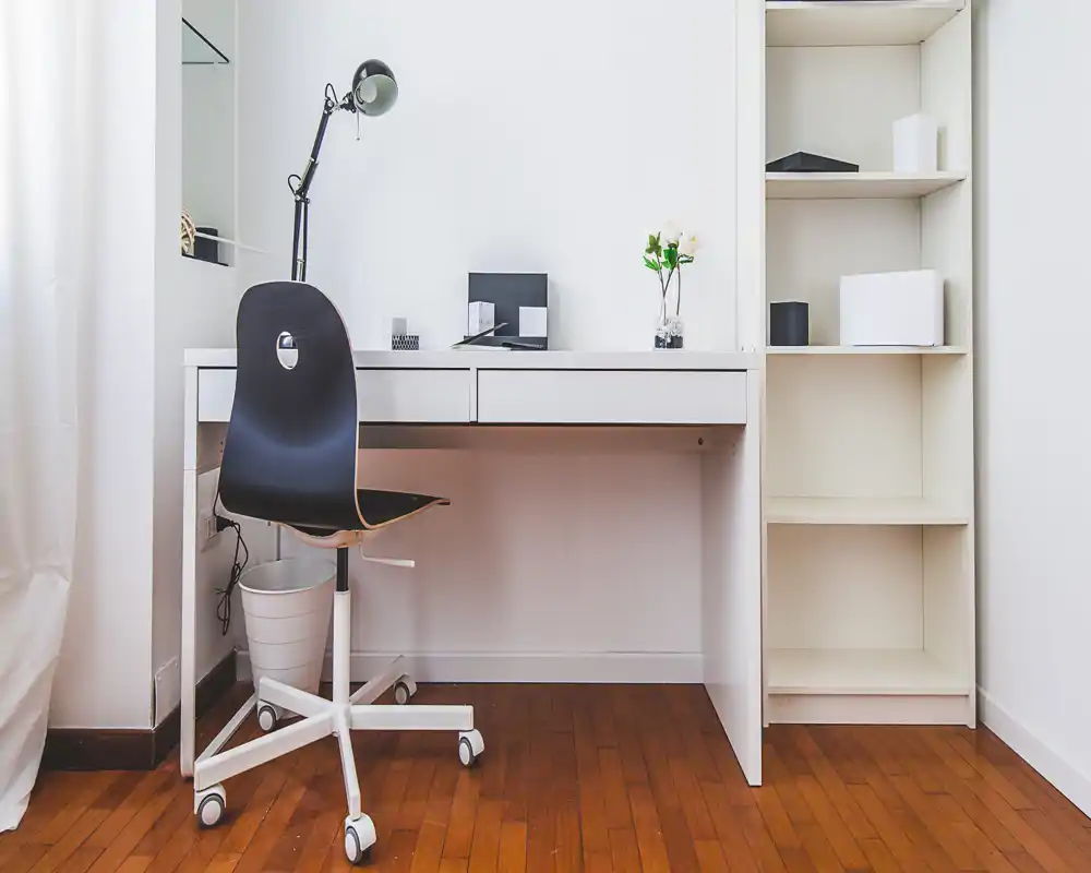 Bright, minimal home office corner with a white desk, shelving and a black swivel chair — neat and well lit for work or study.