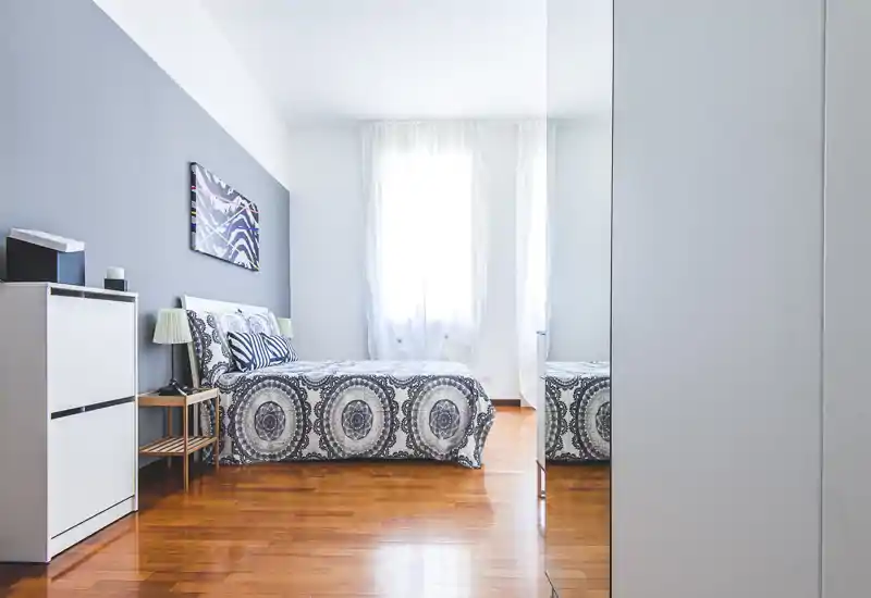 Bright, modern bedroom with patterned bedding, natural light from a large window and a mirrored wardrobe that adds depth. Clean, minimalist styling with wooden floors and decorative cushions.