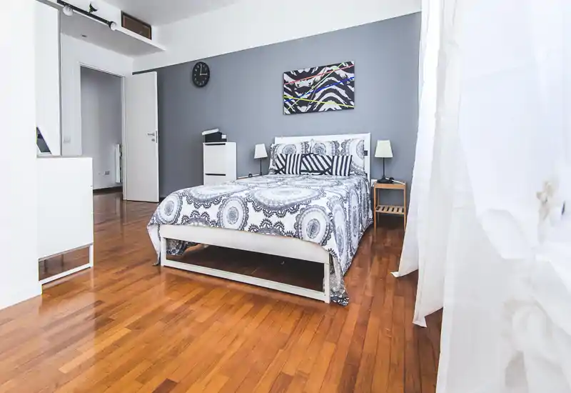 Bright, modern bedroom with a patterned double bed, bedside tables and natural light from a large window. The room features warm wooden floors and minimalist décor for a comfortable stay.