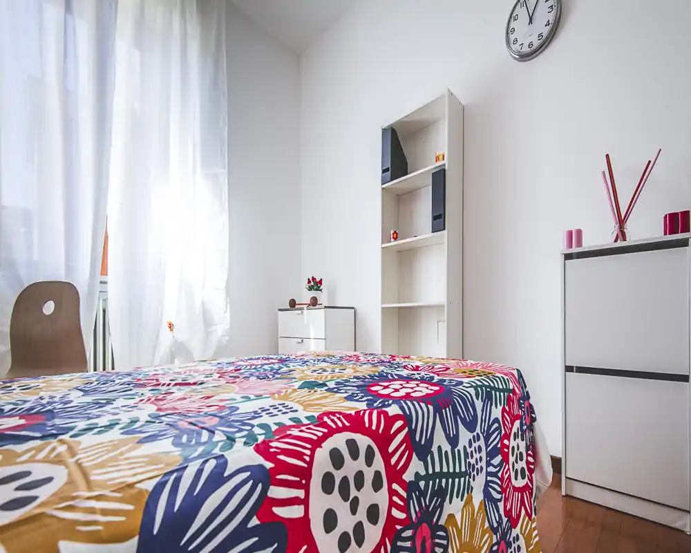 Bright, colorful bedroom with a large bed in the foreground, natural light from a sheer-curtained window and simple modern storage furniture.