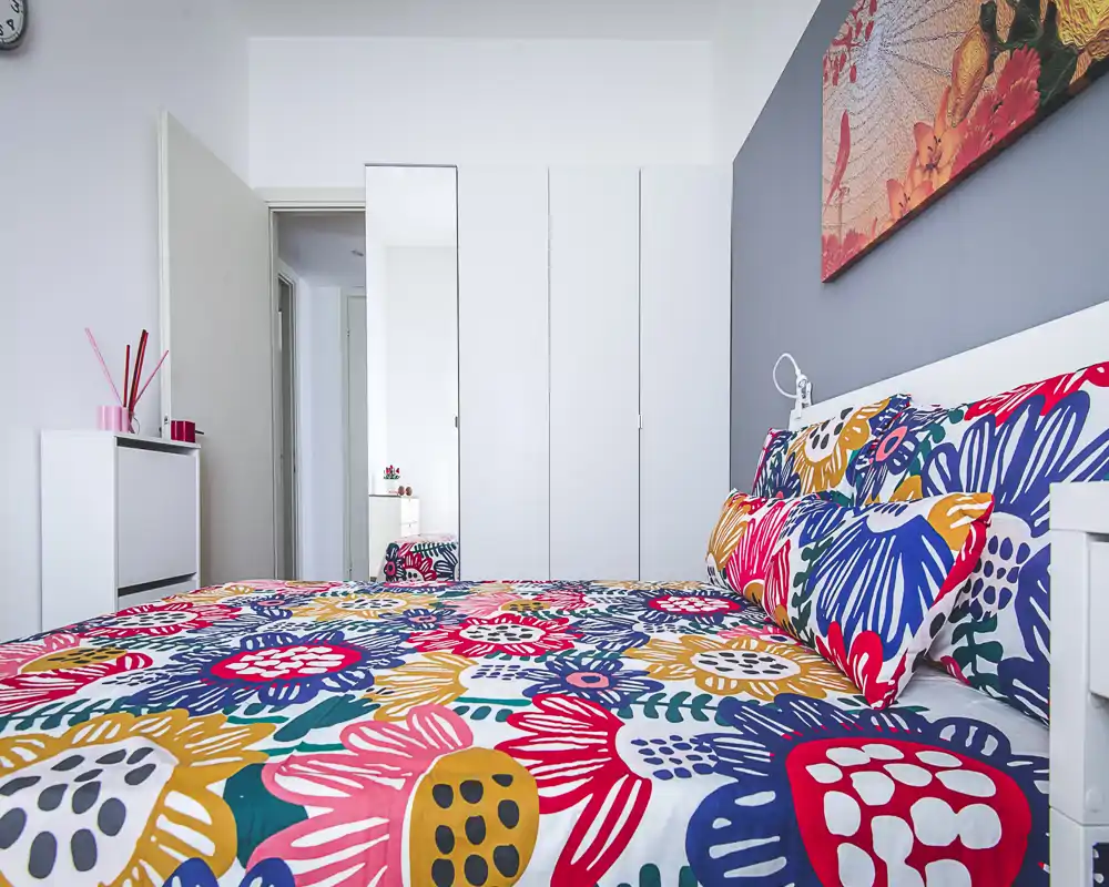 Bright, colorful bedroom with a large floral-patterned bed, coordinated pillows and modern white storage. The room feels fresh and inviting with clean lines and decorative artwork above the headboard.