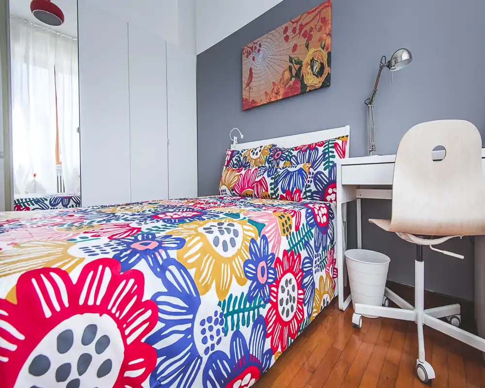 Bright, colorful bedroom with a full visible bed dressed in bold floral bedding, a small desk and chair beside it, and cheerful wall art.