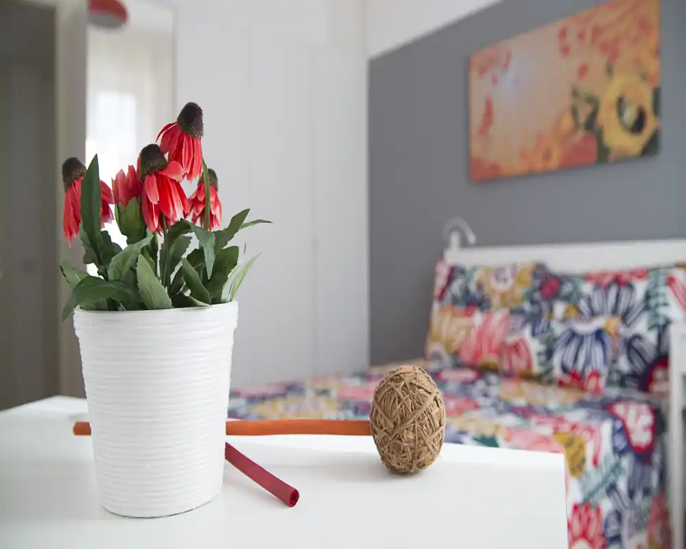 A bright, staged bedroom with colorful patterned bedding and a decorative plant in the foreground; clean modern headboard and artwork create a cheerful, welcoming atmosphere.