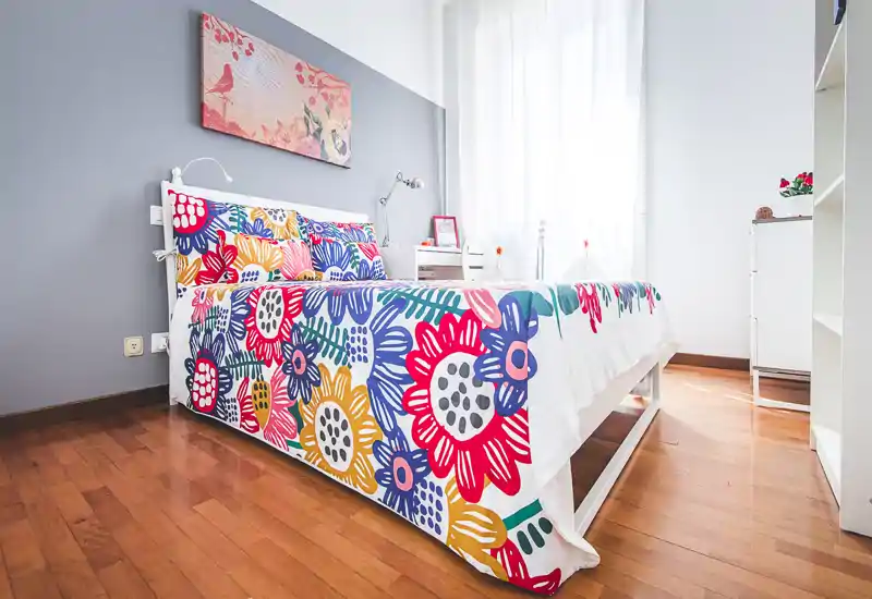 Bright, well-staged bedroom with a colorful floral bedspread, natural light from a large window, and modern minimal furnishings.