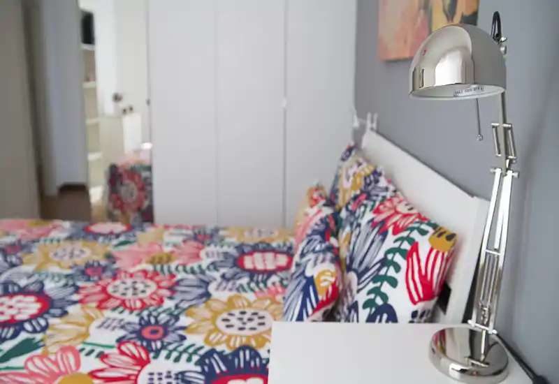 Bright and colorful bedroom featuring a patterned double bed with decorative pillows and a chrome bedside lamp; calm grey walls and a wardrobe in the background create a tidy, stylish space ideal for rentals.