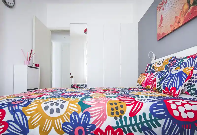 Bright bedroom with a large, colorful bedspread dominating the foreground; white wardrobe and minimal, modern furnishings create a fresh, airy feel.