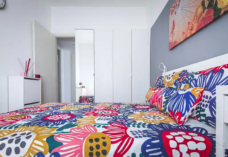 Bright, colorful bedroom with a large floral-patterned bed, coordinated pillows and modern white storage. The room feels fresh and inviting with clean lines and decorative artwork above the headboard.